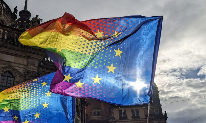 Zu sehen zwei gleiche im Wind wehende Fahnen, die zweigeteilt sind. Links ist die Queer-Pride-Fahne und rechts die Europa-Fahne abgebildet. Die Fahnen schwingen vor dem Schoss Dresden